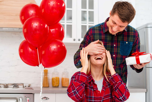 Un homme surprend sa partenaire avec un cadeau dans la cuisine, entouré de ballons rouges, tous deux souriants
